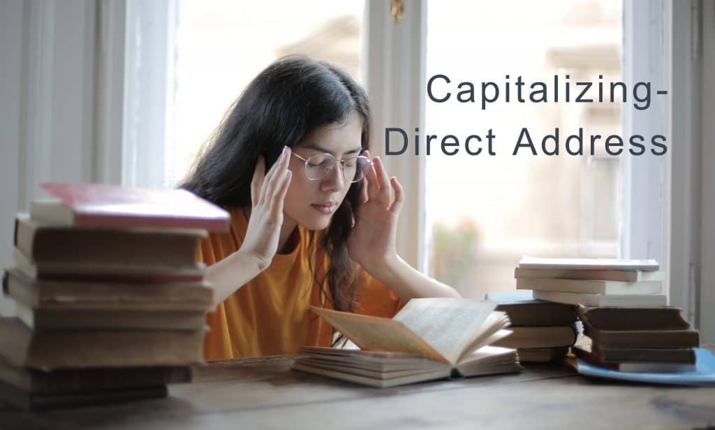young woman sitting at a table studying surrounded by books. She is wearing glasses and pressing her fingers to her temples as if she has a headache.  text in the photo says " Capitalizing- Direct Address"